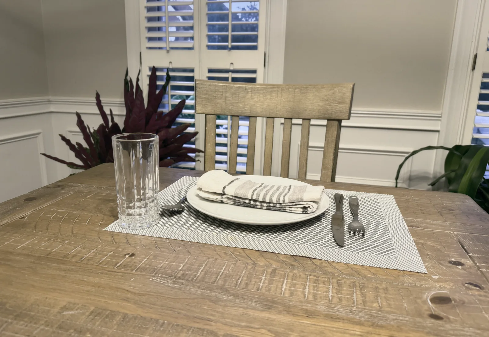 A place set at the wooden dining table beside shuttered windows.